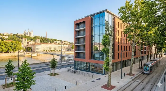 Une photographie extérieure lumineuse de jour capturant la façade d'une résidence étudiante moderne à Lyon 7ème, située le long du Rhône. Le bâtiment contemporain présente un mélange de briques et de verre, intégré à la rue avec une ligne de tramway TCL TCL B et des arbres. À l'arrière-plan, au-delà du Rhône, une vue panoramique de Lyon est visible, incluant la distant Basilique de Fourvière sur sa colline et la Tour Métallique de Fourvière sous un ciel bleu clair. Il n'y a aucun texte sur l'image.