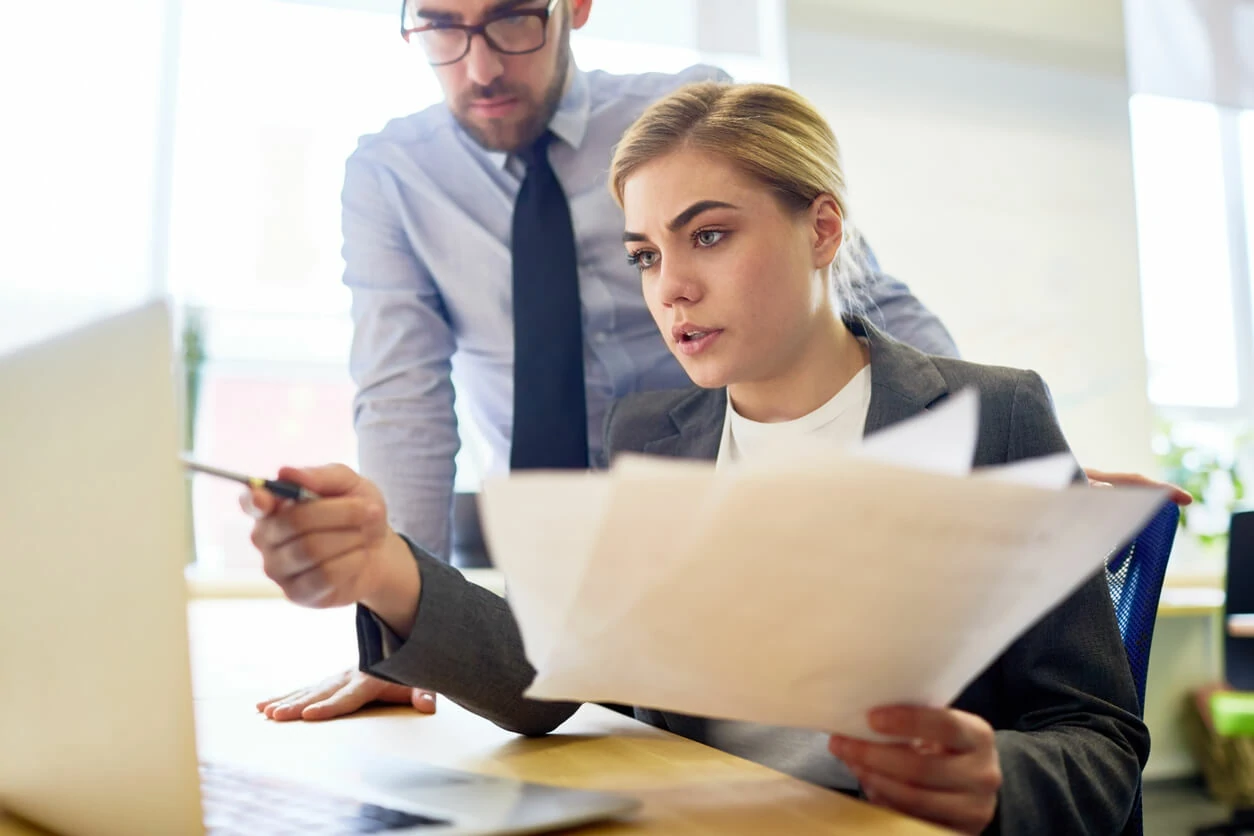 Un homme debout derrière une femme qui tient des documents et regardant dans un ordinateur portable.