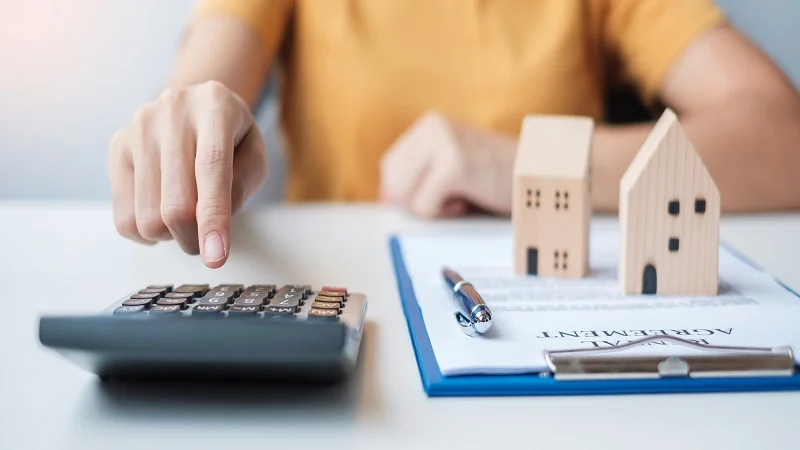 Main utilisant une calculatrice à côté de petites maisons en bois et des documents